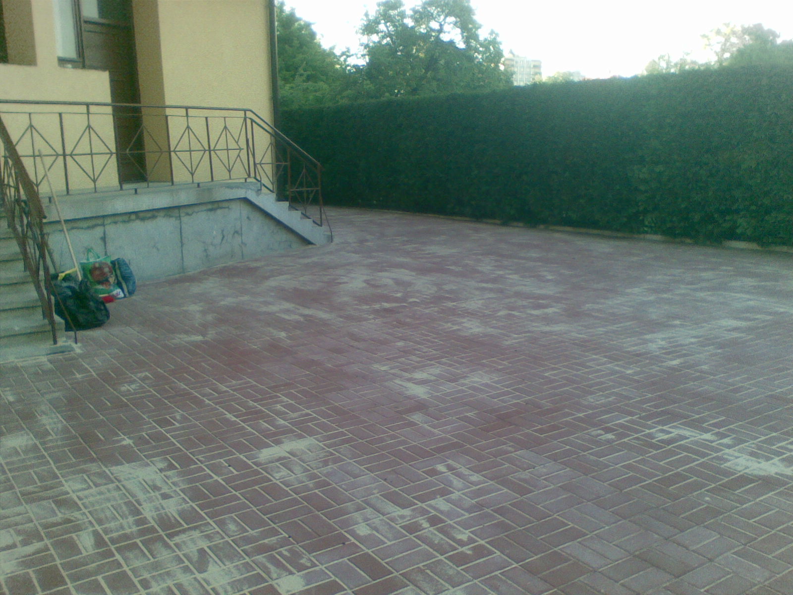 Ułożona kostka brukowa w jodełkę na podjeździe przed domem z zielonym żywopłotem w tle, widoczne schody wejściowe z metalową barierką.