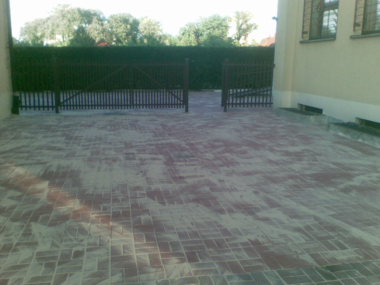 Ułożona kostka brukowa w odcieniach czerwieni i szarości przed posesją z drewnianą bramą i zielonym żywopłotem w tle.