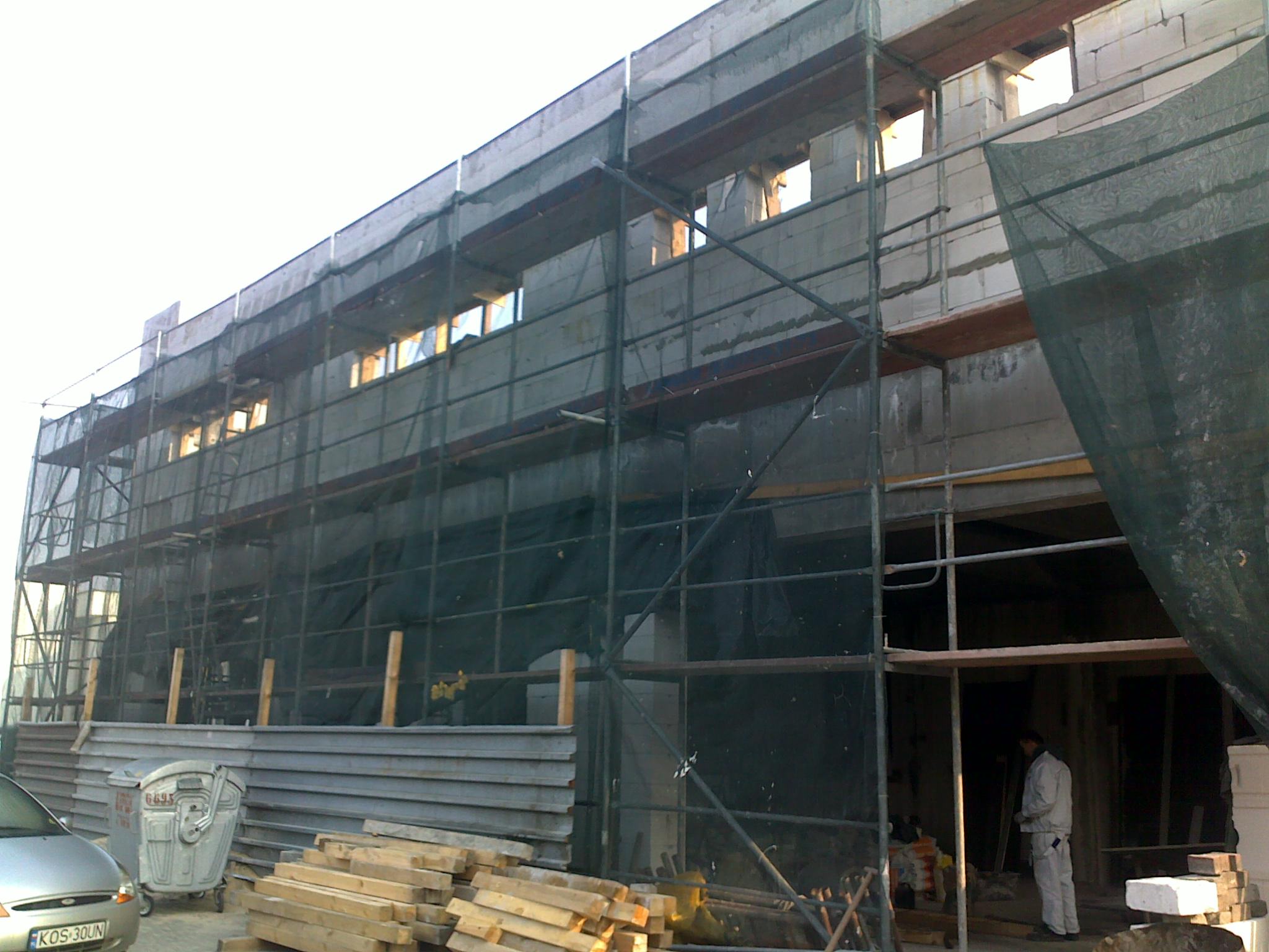 Wznoszony budynek przemysłowy z białego bloczka, osłonięty siatką na rusztowaniach, widoczny pracownik wewnątrz, obok składowane materiały budowlane i kontener na śmieci.