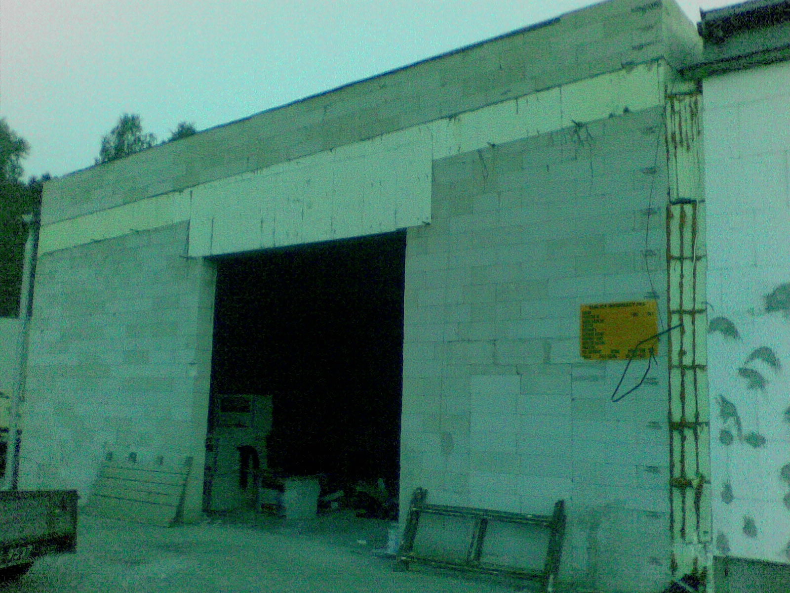 Budynek przemysłowy w trakcie budowy z pustaków, widoczne elementy konstrukcyjne, ślady po zaprawie i żółta tablica informacyjna na ścianie