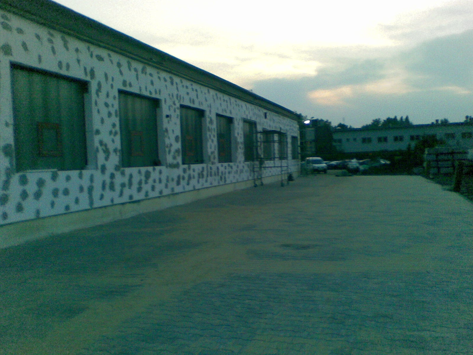 Budynek z elewacją w trakcie malowania, widoczne plamy niedomalowanej farby, rusztowanie przy oknie, kostka brukowa na placu przed budynkiem.