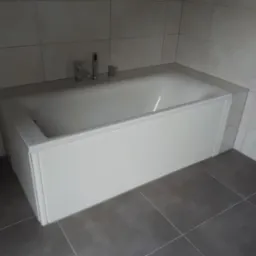 Zabudowana wanna z białą obudową i chromowaną armaturą w łazience wyłożonej szarymi płytkami.