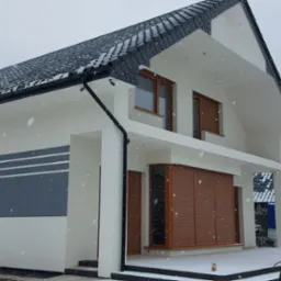 Nowoczesny dom jednorodzinny w Wągrowcu po ociepleniu elewacji w zimowej scenerii, widoczne białe ściany z szarymi pasami, brązowe okna z roletami i lekki śnieg.