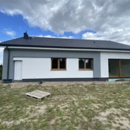 Świeżo otynkowany, parterowy dom jednorodzinny z szarą i białą elewacją, widoczne drewniane okna i przesuwne drzwi balkonowe, paleta na trawniku, błękitne niebo z chmurami w tle.