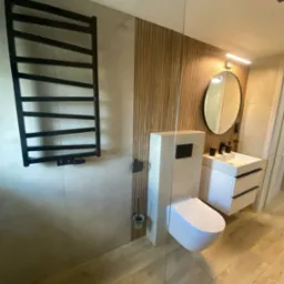 Nowoczesna łazienka z drewnianymi lamelami, owalnym lustrem, białymi szafkami i podwieszaną toaletą. Czarny grzejnik drabinkowy i drewniana podłoga.