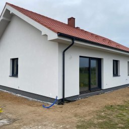 Z.U.P.H. Wojciech Zdrzałka i synowie - Nowoczesny dom jednorodzinny z białą elewacją, czerwoną dachówką i czarnymi oknami. Detal: niebieski wąż przy fundamencie. Widok z zewnątrz, pochmurne niebo.