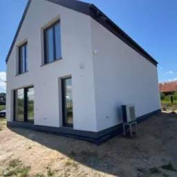 Nowoczesny dom z białą elewacją i czarnymi ramami okiennymi, widziany z zewnątrz w słoneczny dzień. Widoczny klimatyzator na zewnątrz i elementy wykończenia.