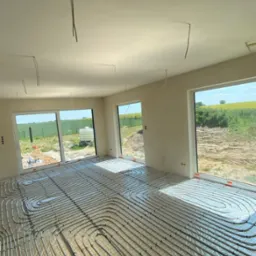 Widok na ułożone rury ogrzewania podłogowego w jasnym pomieszczeniu z dużymi oknami, za którymi widać zielone pola i błękitne niebo. W tle surowe ściany.