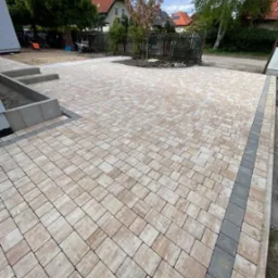 Ułożona kostka brukowa w odcieniach beżu i szarości, tworząca wzór na posesji. Widoczne obrzeża i fragmenty zieleni w tle, w słoneczny dzień.