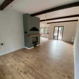 Z.U.P.H. Wojciech Zdrzałka i synowie - Jasne, przestronne wnętrze po remoncie: drewniana podłoga, szary kominek z drewnianą półką, białe ściany i ciemne belki stropowe. Widok na okno i drzwi balkonowe.