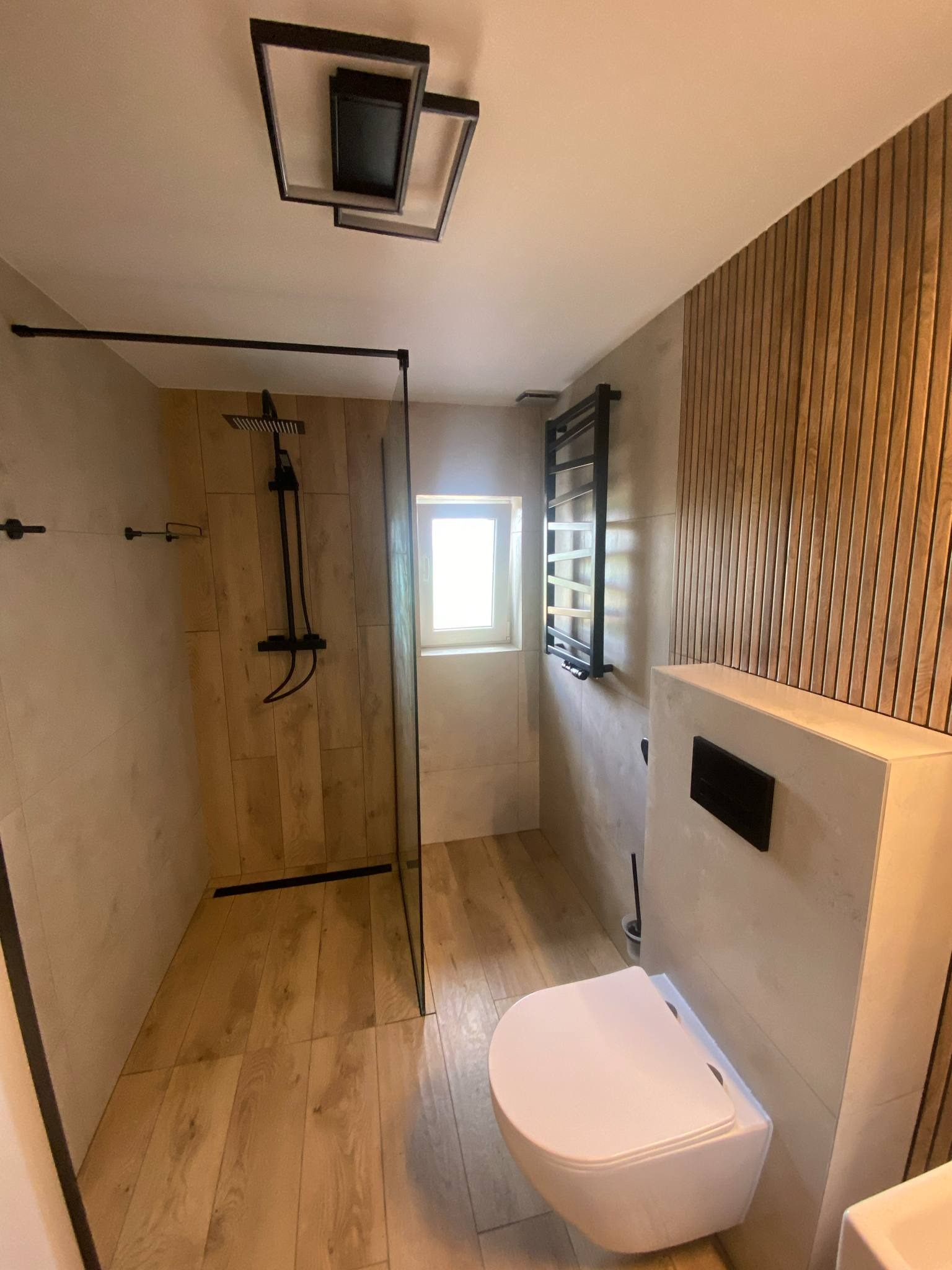 Nowoczesna łazienka z kabiną typu walk-in, drewnianą podłogą i ścianą z paneli. Czarna armatura i grzejnik drabinkowy dodają elegancji. Minimalistyczne oświetlenie.