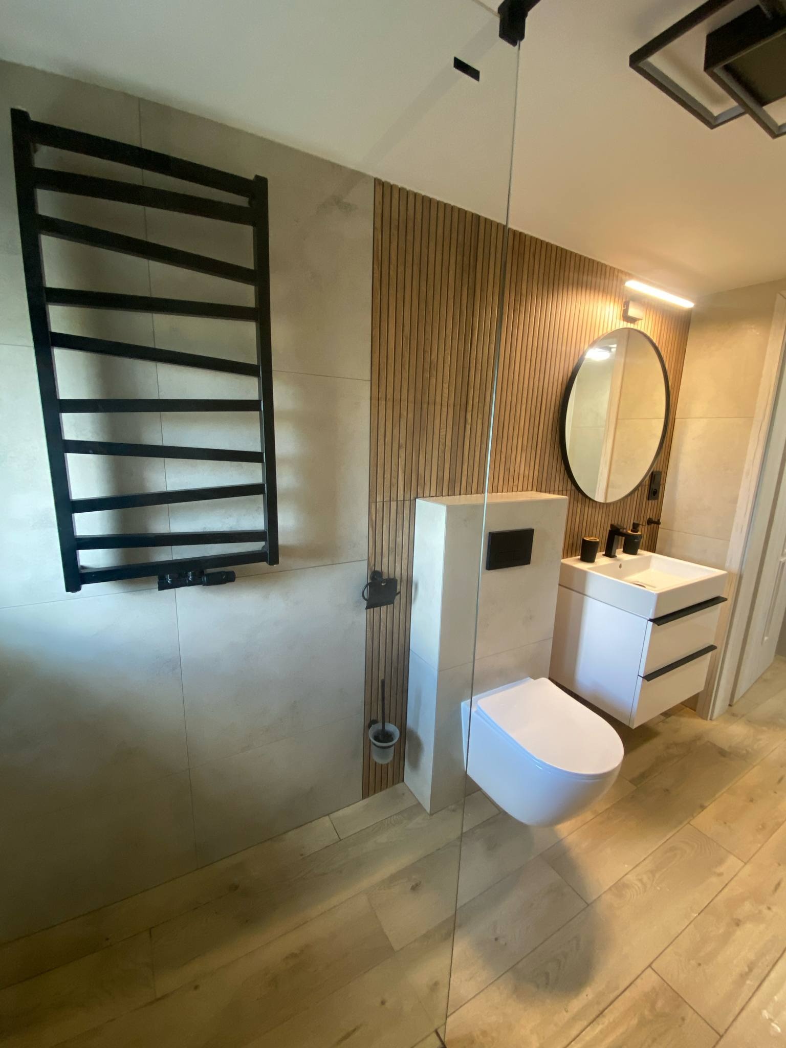 Nowoczesna łazienka z drewnianymi lamelami, owalnym lustrem, białymi szafkami i podwieszaną toaletą. Czarny grzejnik drabinkowy i drewniana podłoga.