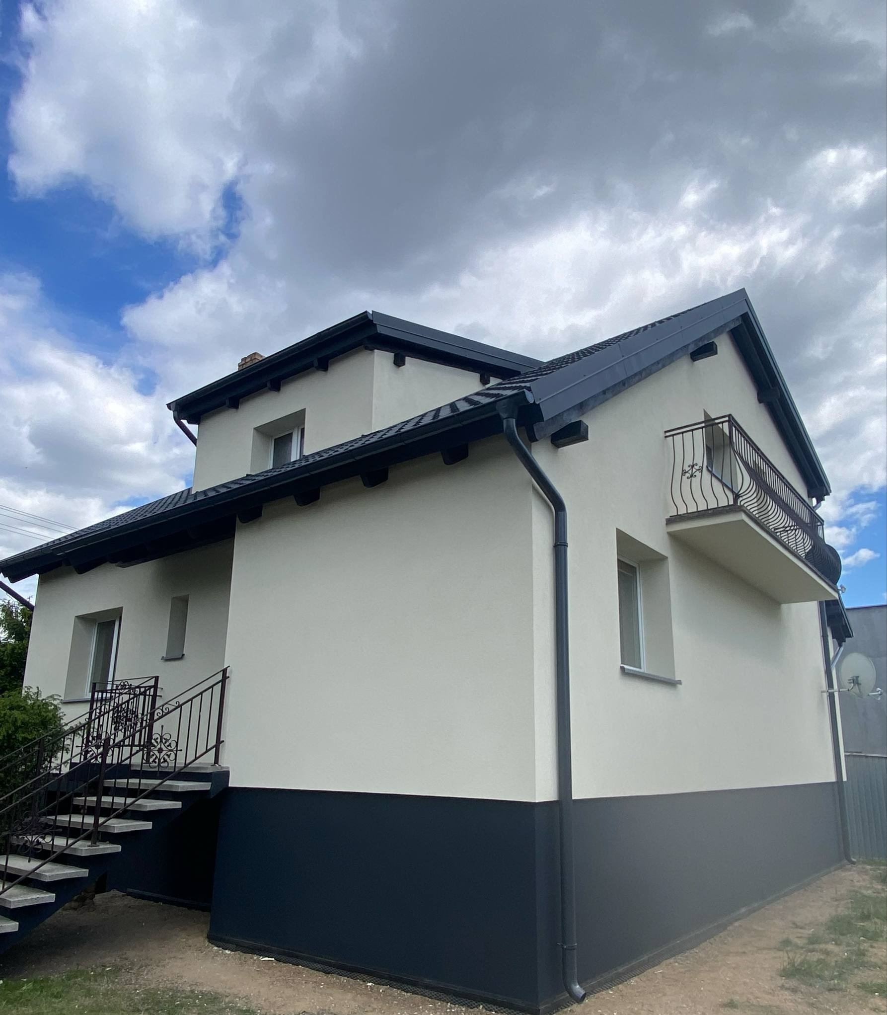 Dwukondygnacyjny dom z balkonem i schodami zewnętrznymi, świeżo po remoncie elewacji, w odcieniach bieli i grafitu na tle pochmurnego nieba.