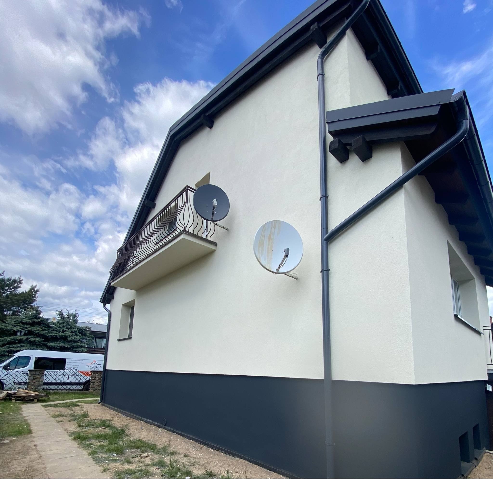 Dwukolorowa elewacja domu z balkonem i dwiema antenami satelitarnymi. Ciemna podmurówka, jasna góra. Widok z dołu, błękitne niebo.