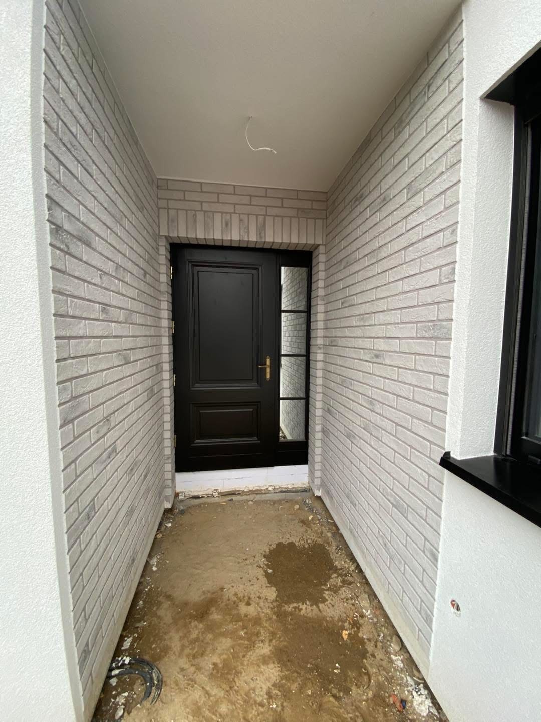 Wejście do domu z czarnymi drzwiami i elewacją z szarej cegły. Widoczny fragment okna. Surowy stan podłoża przed wykończeniem.
