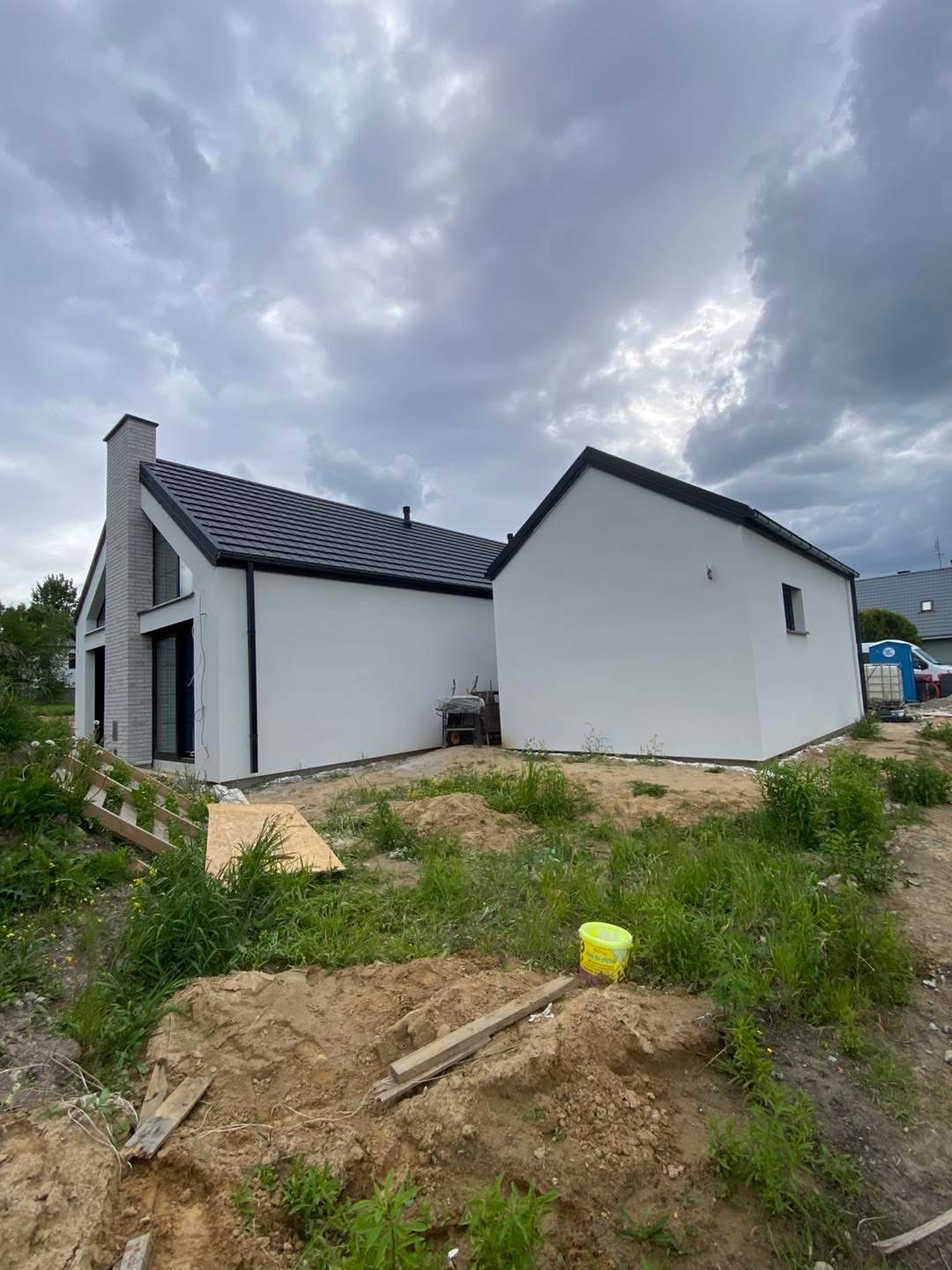 Biały dom z ciemną dachówką pod pochmurnym niebem, widok z poziomu gruntu. Wokół domu widoczne pozostałości po budowie, kubeł farby i narzędzia.