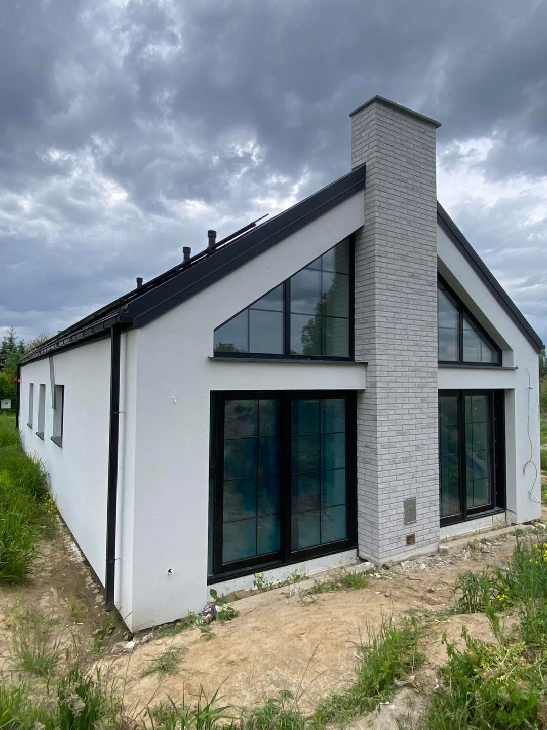 Nowoczesny dom z białą elewacją, czarnymi oknami i ceglanym kominem na tle pochmurnego nieba. Widoczne trawy i ziemia wokół budynku, co sugeruje świeżo zakończone prace.