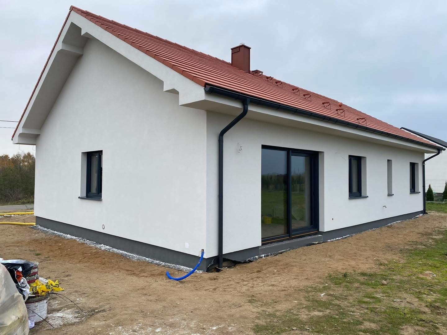 Nowoczesny dom jednorodzinny z białą elewacją, czerwoną dachówką i czarnymi oknami. Detal: niebieski wąż przy fundamencie. Widok z zewnątrz, pochmurne niebo.