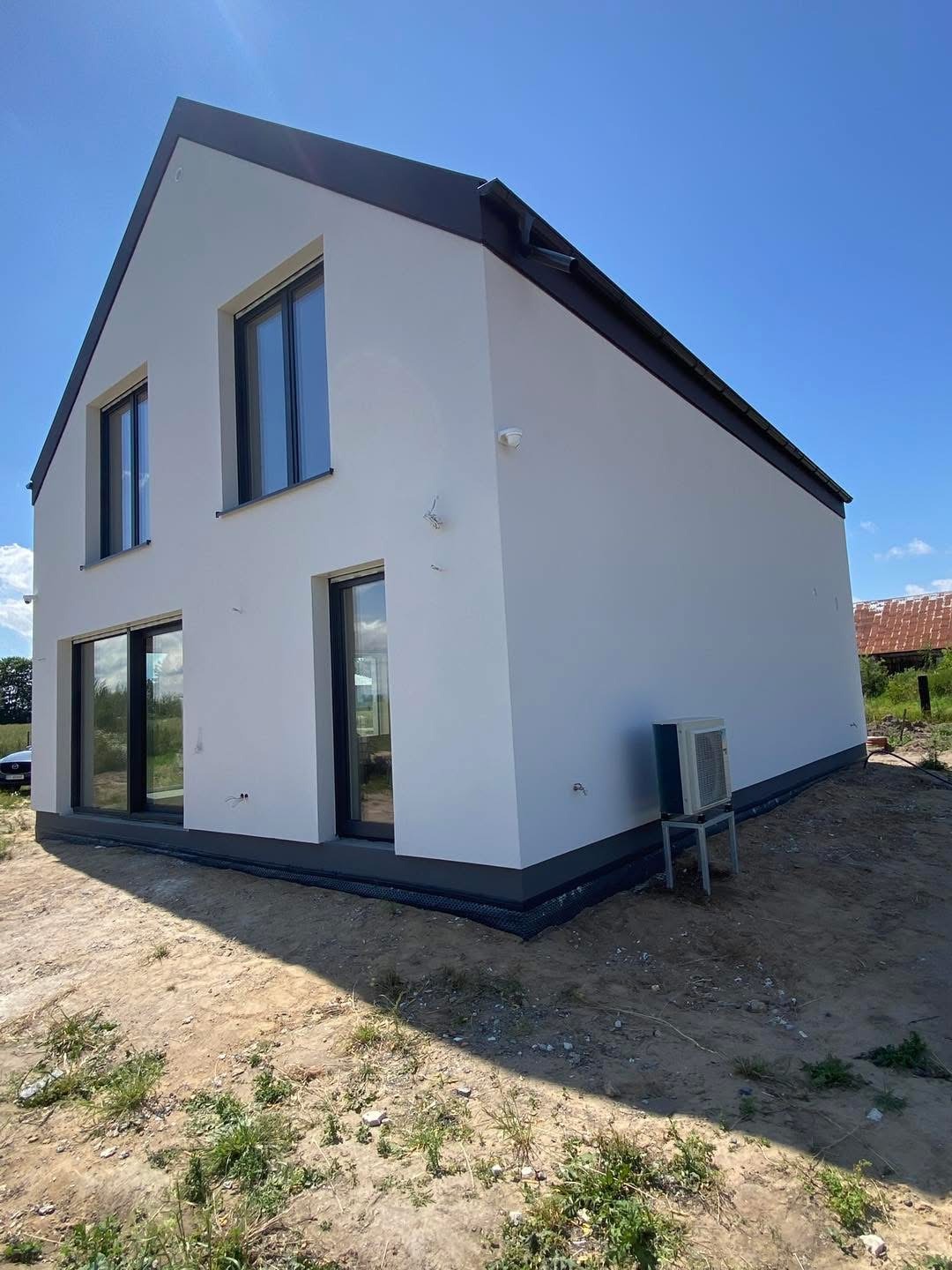 Nowoczesny dom z białą elewacją i czarnymi ramami okiennymi, widziany z zewnątrz w słoneczny dzień. Widoczny klimatyzator na zewnątrz i elementy wykończenia.