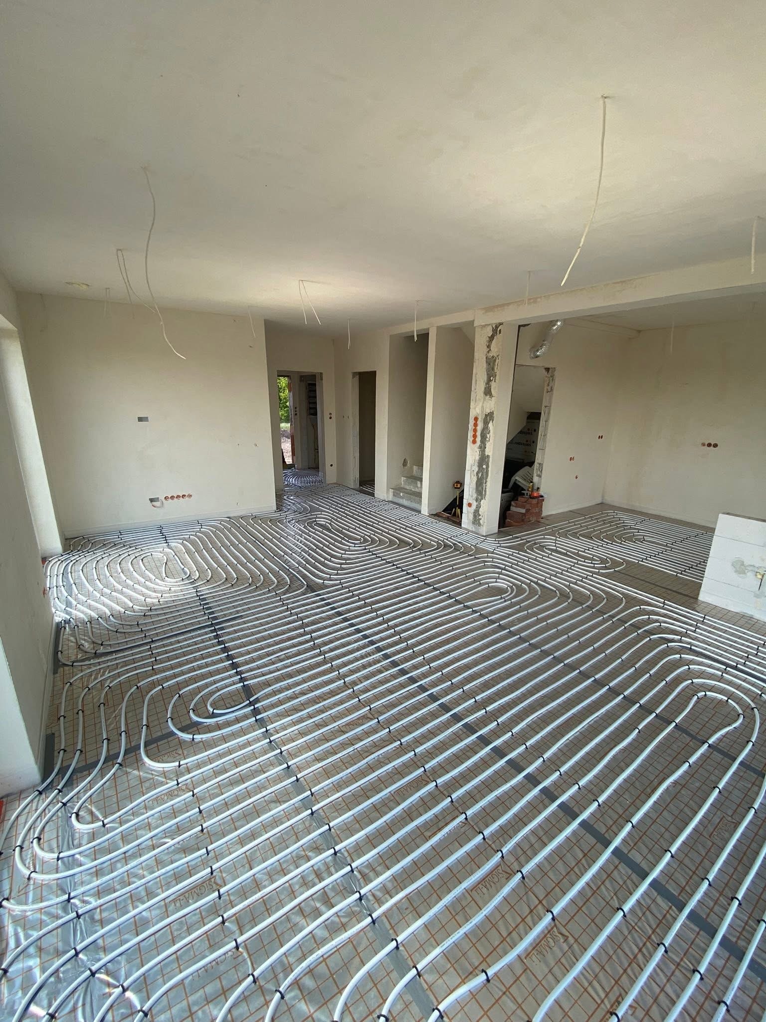 Białe rury ogrzewania podłogowego ułożone spiralnie na folii termoizolacyjnej w surowym wnętrzu z widocznymi instalacjami elektrycznymi w ścianach.