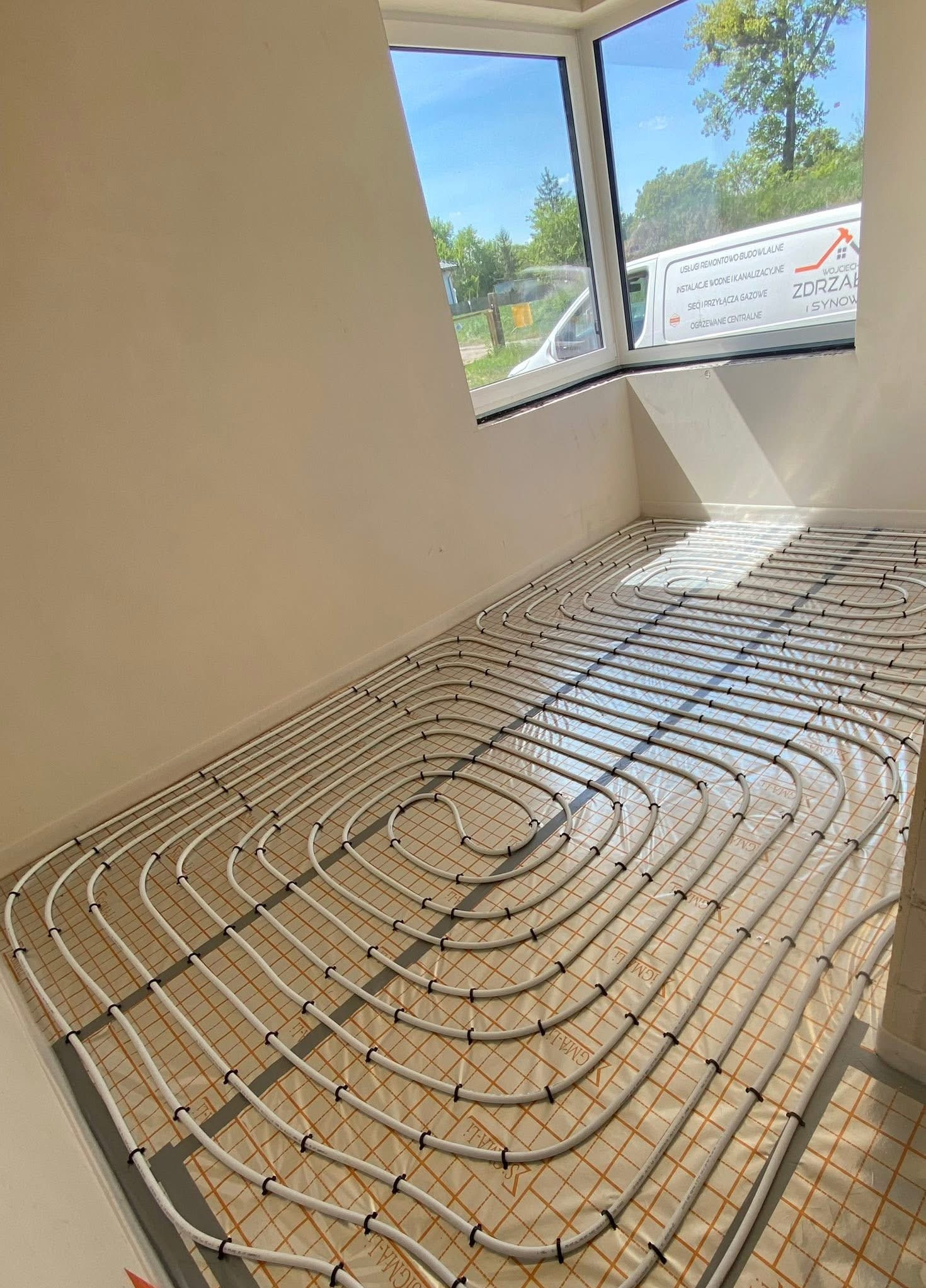 Białe rury ogrzewania podłogowego ułożone spiralnie na pomarańczowej macie w jasnym pokoju z oknem, przez które widać zieleń. Widoczny fragment busa firmy.