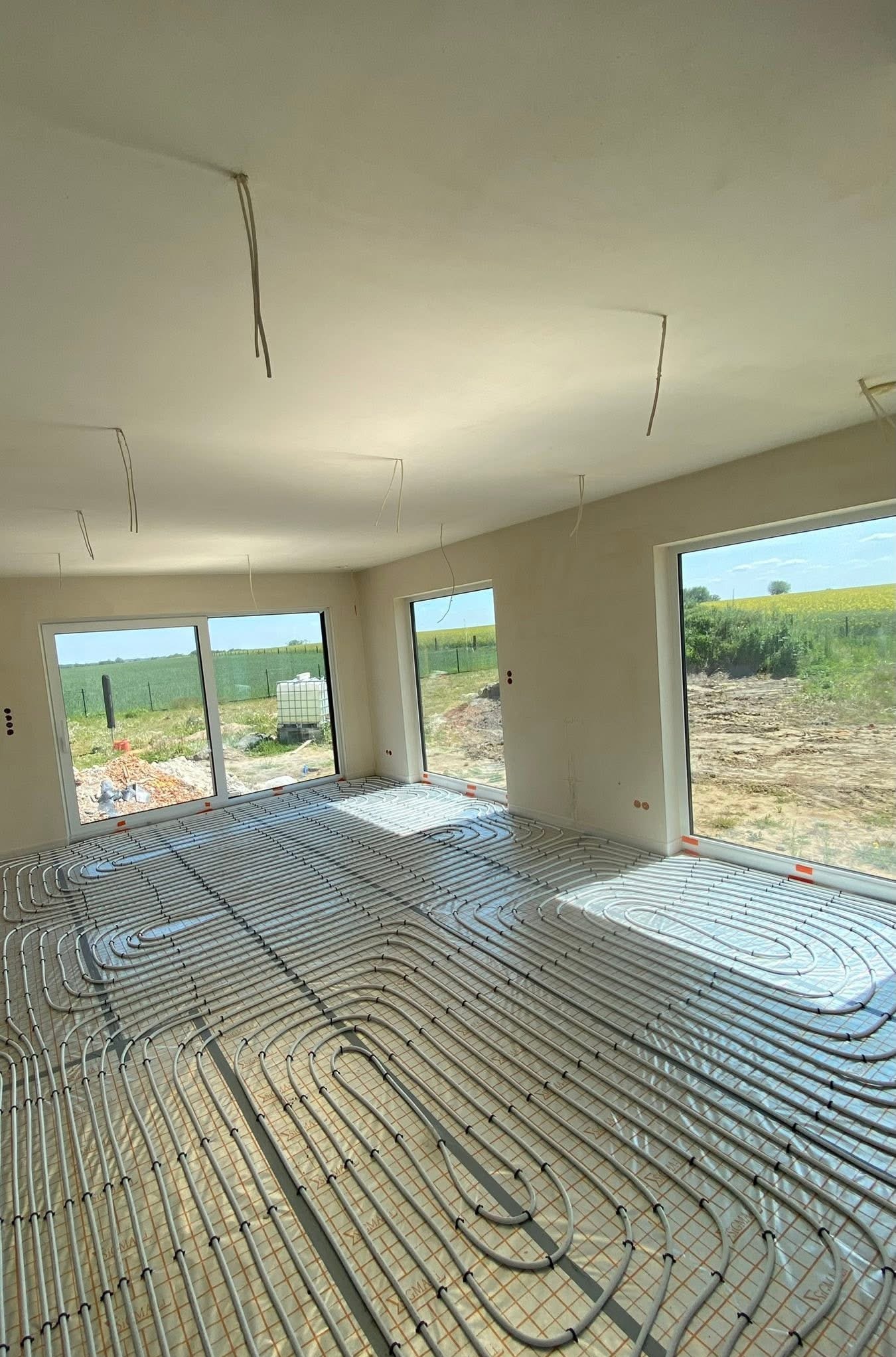 Widok na ułożone rury ogrzewania podłogowego w jasnym pomieszczeniu z dużymi oknami, za którymi widać zielone pola i błękitne niebo. W tle surowe ściany.