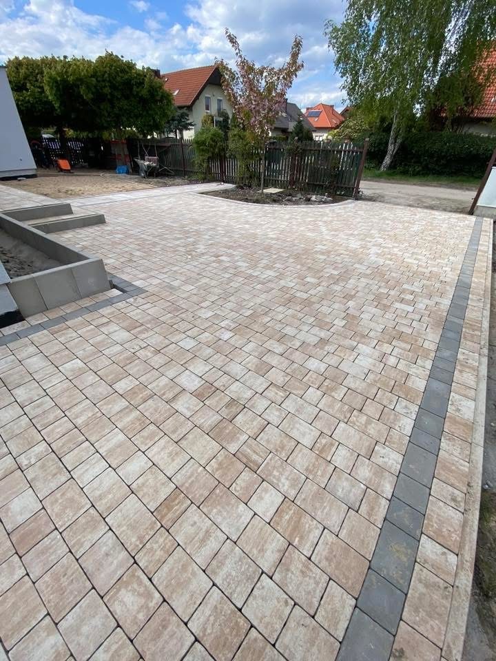 Ułożona kostka brukowa w odcieniach beżu i szarości, tworząca wzór na posesji. Widoczne obrzeża i fragmenty zieleni w tle, w słoneczny dzień.