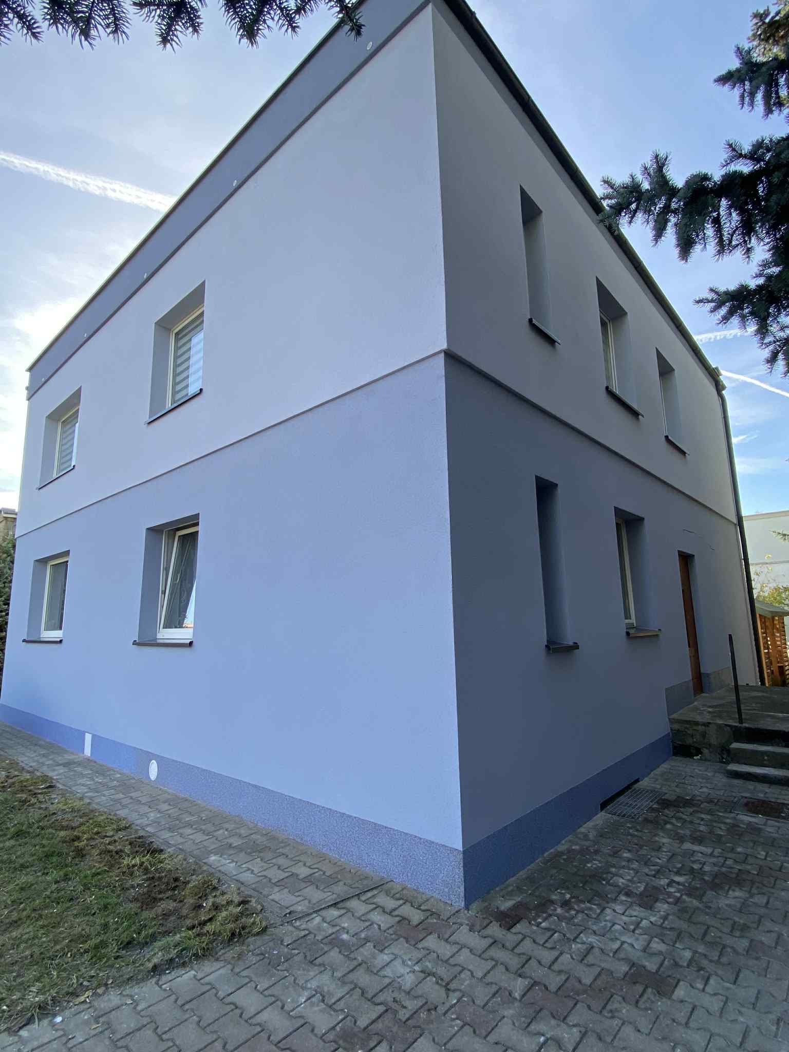 Dwukondygnacyjny dom po remoncie elewacji, pomalowany na dwa odcienie szarości, z widocznymi oknami i fragmentem chodnika z kostki brukowej. Ujęcie z dołu.