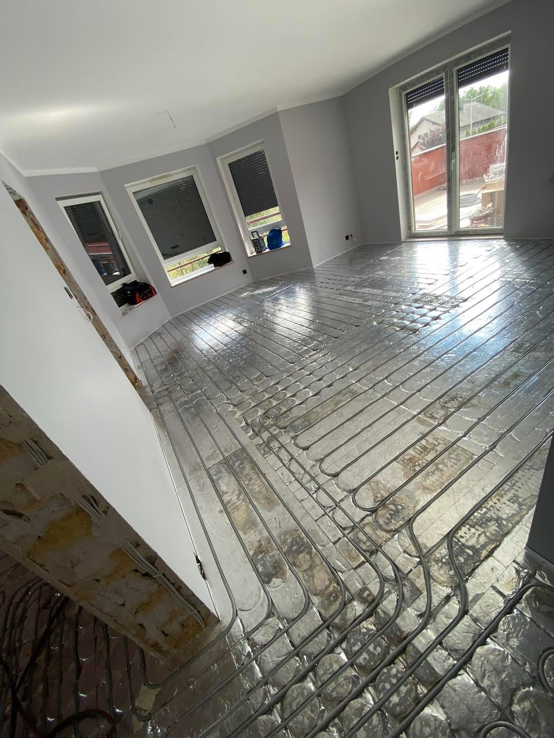 Szare wnętrze z ułożonym systemem ogrzewania podłogowego na folii aluminiowej, widoczne okna i drzwi balkonowe, przygotowane do wylewki.