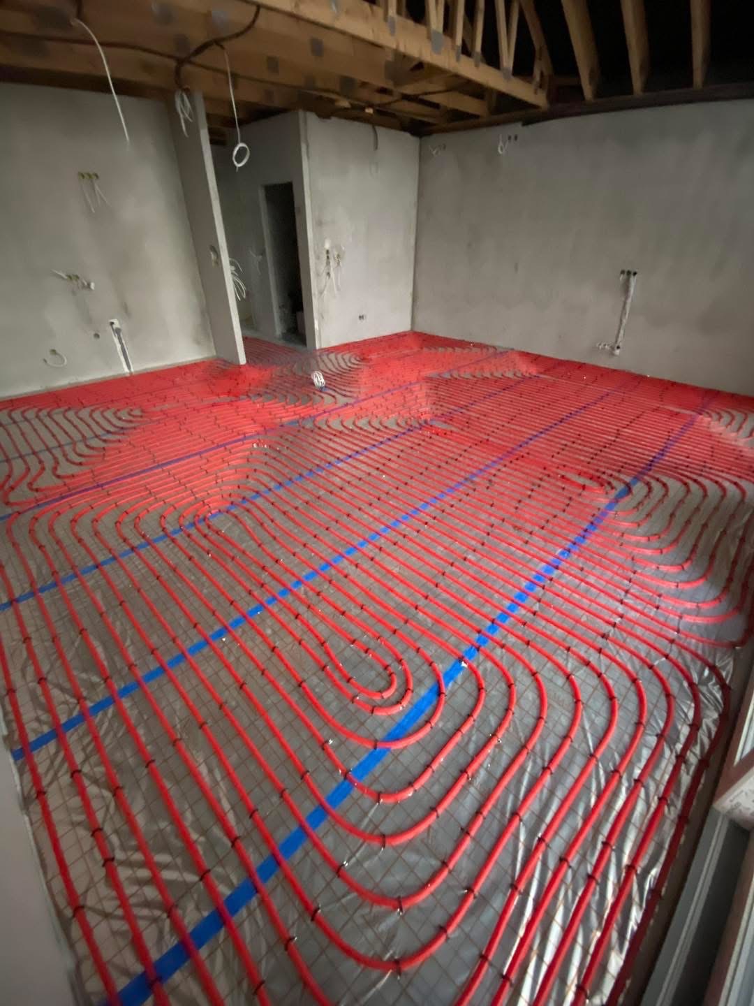 Czerwone rury ogrzewania podłogowego ułożone na folii w surowym wnętrzu z widoczną konstrukcją dachu. Niebieska taśma mocująca. Etap instalacji przed wylewką.