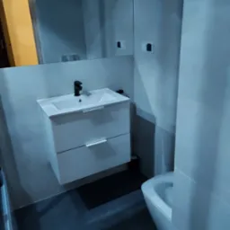 Nowoczesna łazienka z białą szafką pod umywalkę, czarną baterią i dużym lustrem, widok na fragment toalety, szare płytki na ścianach i podłodze.