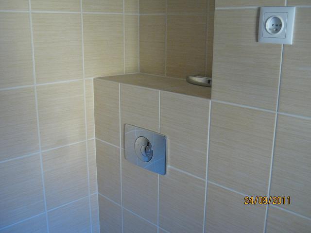 Beżowe płytki ceramiczne na ścianie łazienki, z widocznym przyciskiem spłuczki WC i gniazdkiem elektrycznym, wykończone białą fugą.