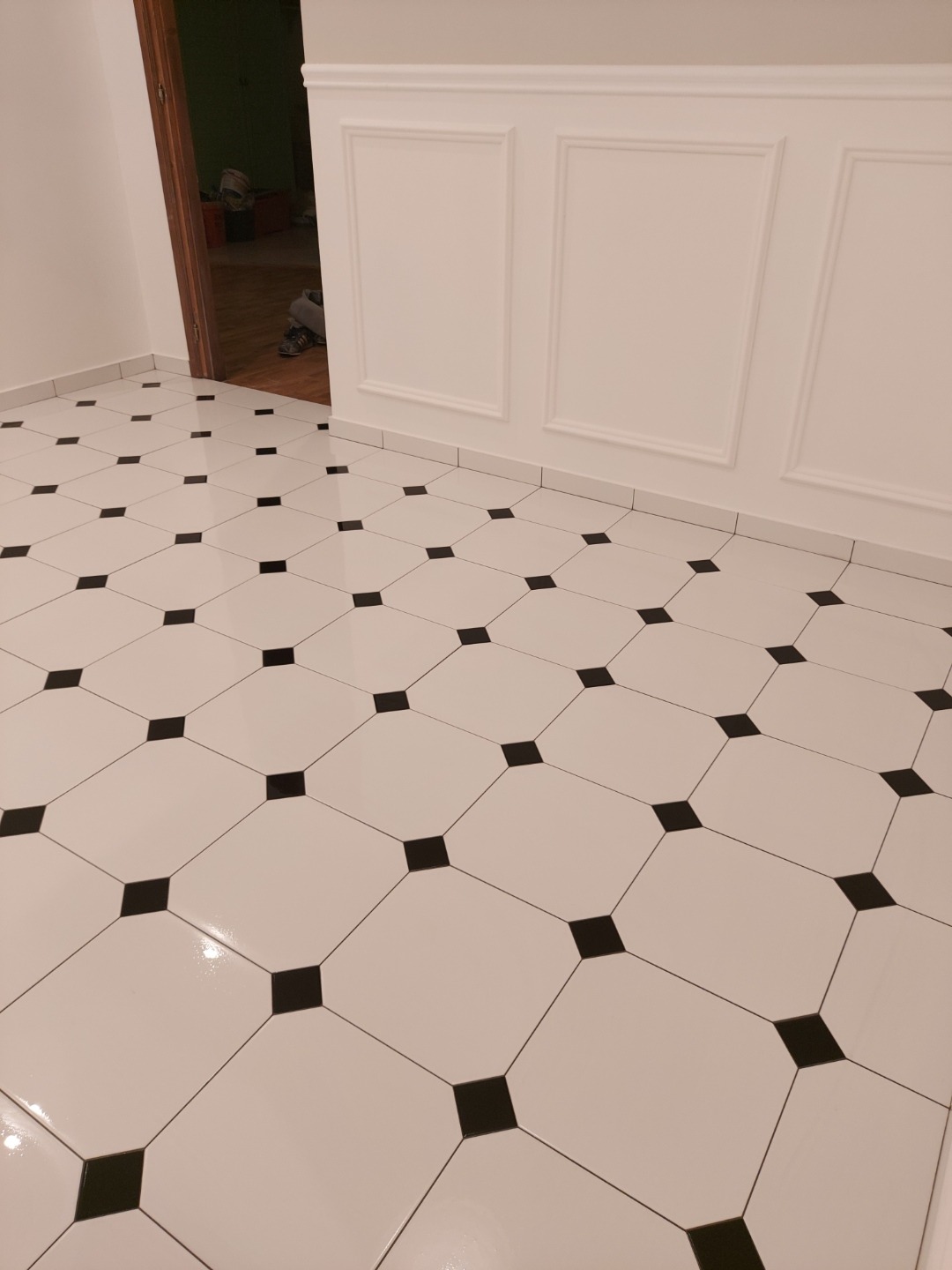 Białe, błyszczące płytki podłogowe z czarnymi, kwadratowymi wstawkami ułożone diagonalnie w nowo wykończonym pomieszczeniu z białymi ścianami i dekoracyjnymi panelami.