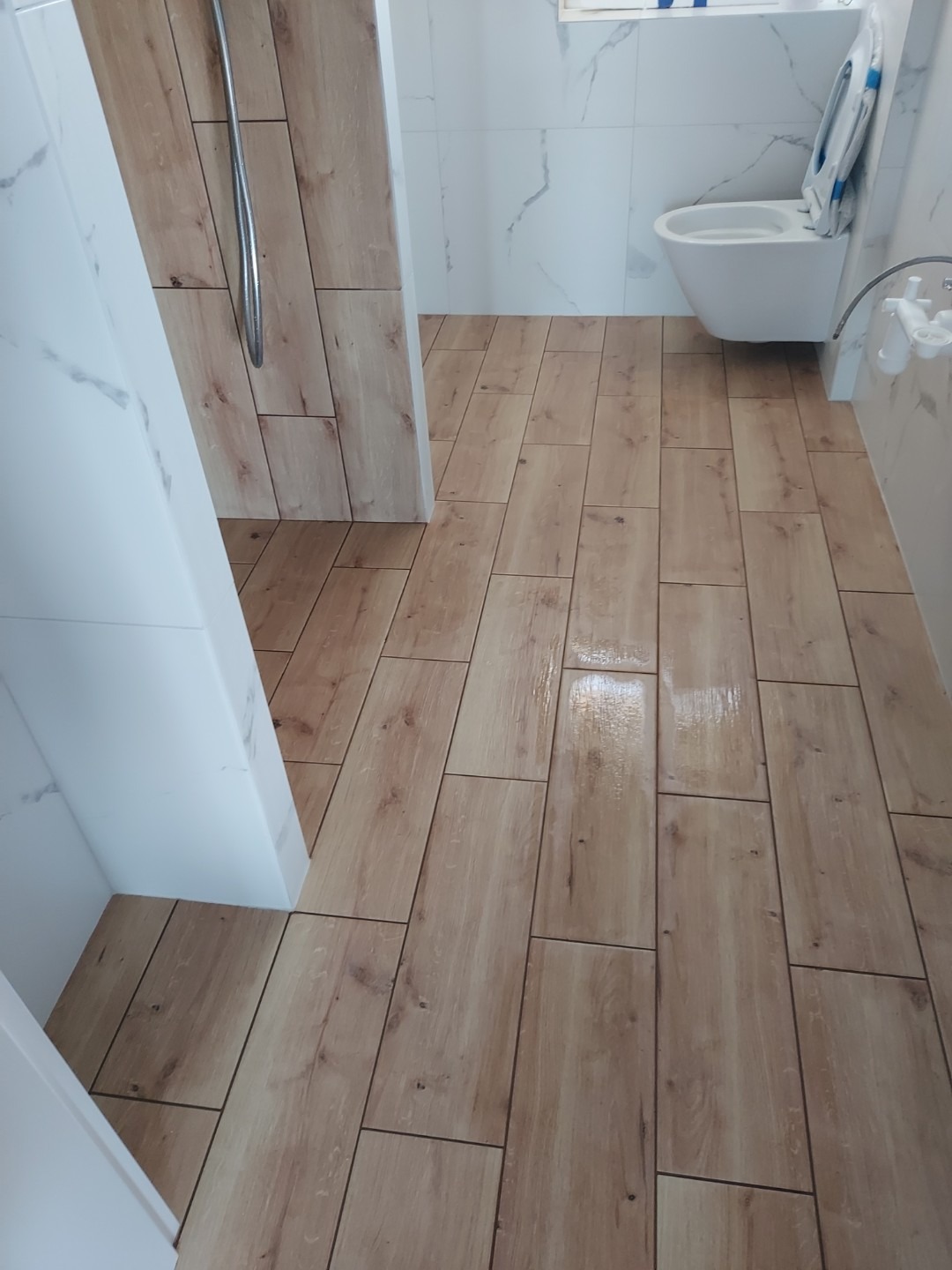 Wykończona łazienka z drewnopodobną podłogą z płytek, białe płytki ścienne z szarymi żyłkami, widoczny fragment kabiny prysznicowej z chromowaną słuchawką, oraz wisząca toaleta.