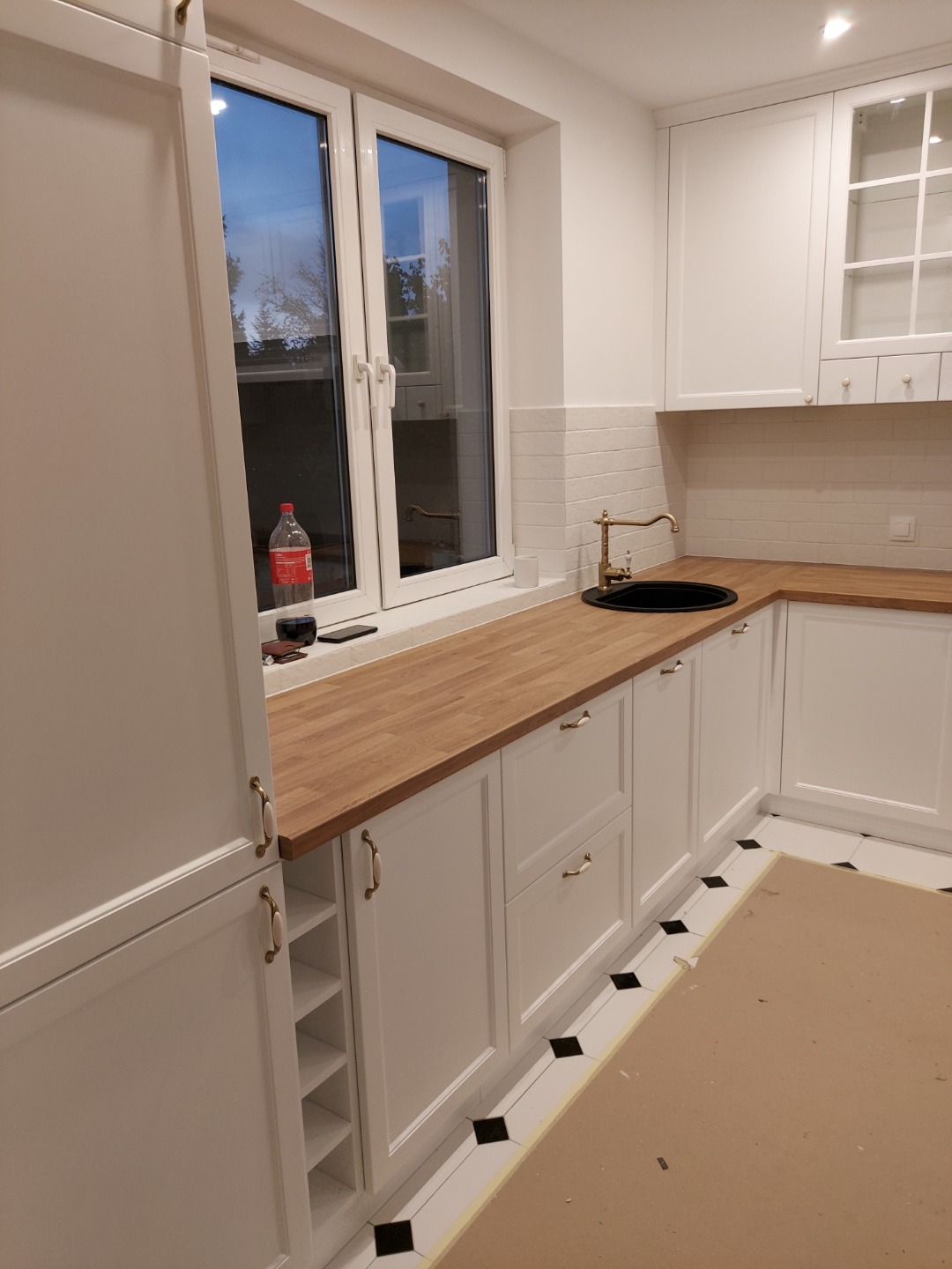 Jasna kuchnia z drewnianym blatem, białymi szafkami z ozdobnymi uchwytami, złotą baterią i czarnym zlewem, widok na okno z butelką Coca-Coli na parapecie, podłoga zabezpieczona kartonem.