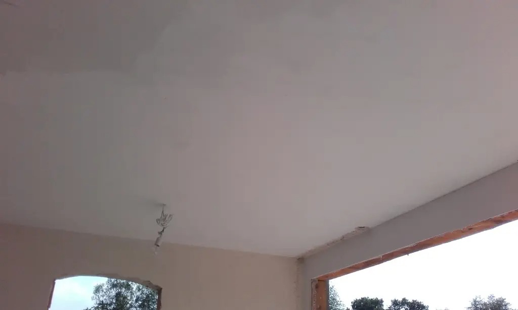 Gładki, jasny sufit po szpachlowaniu z widocznymi przewodami elektrycznymi w miejscu mocowania lampy, fragment ściany z oknem i widokiem na zieleń na zewnątrz.