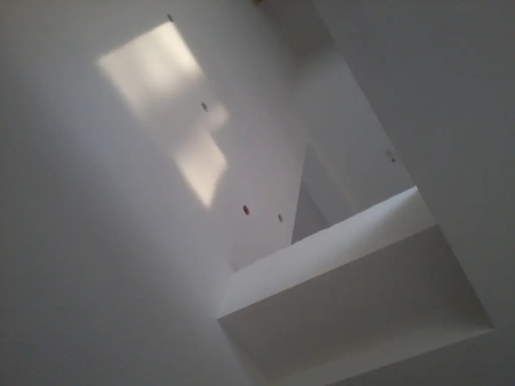Białe ściany i sufit z wykończonym otworem, widoczne ślady po wkrętach, oświetlenie odbija się od gładkiej powierzchni.