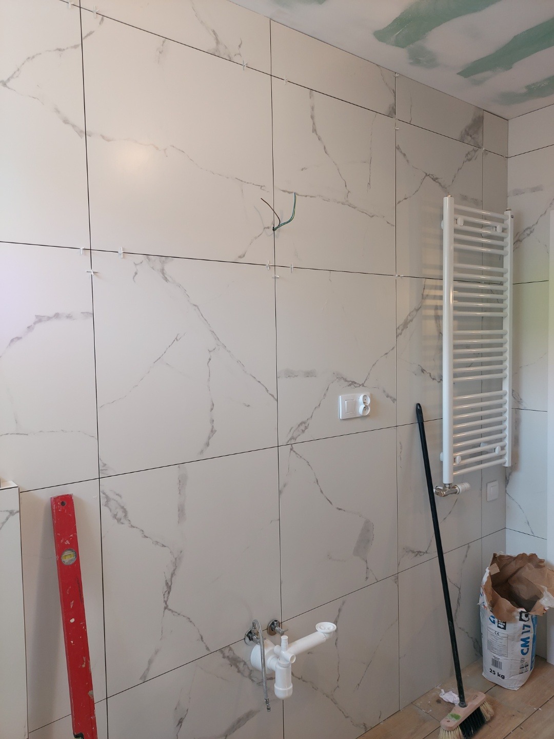 Ściana w łazience wyłożona białymi płytkami imitującymi marmur, z widocznymi krzyżykami dystansowymi, zamontowany grzejnik drabinkowy, poziomica oparta o ścianę, rury odpływowe i workiem zaprawy.