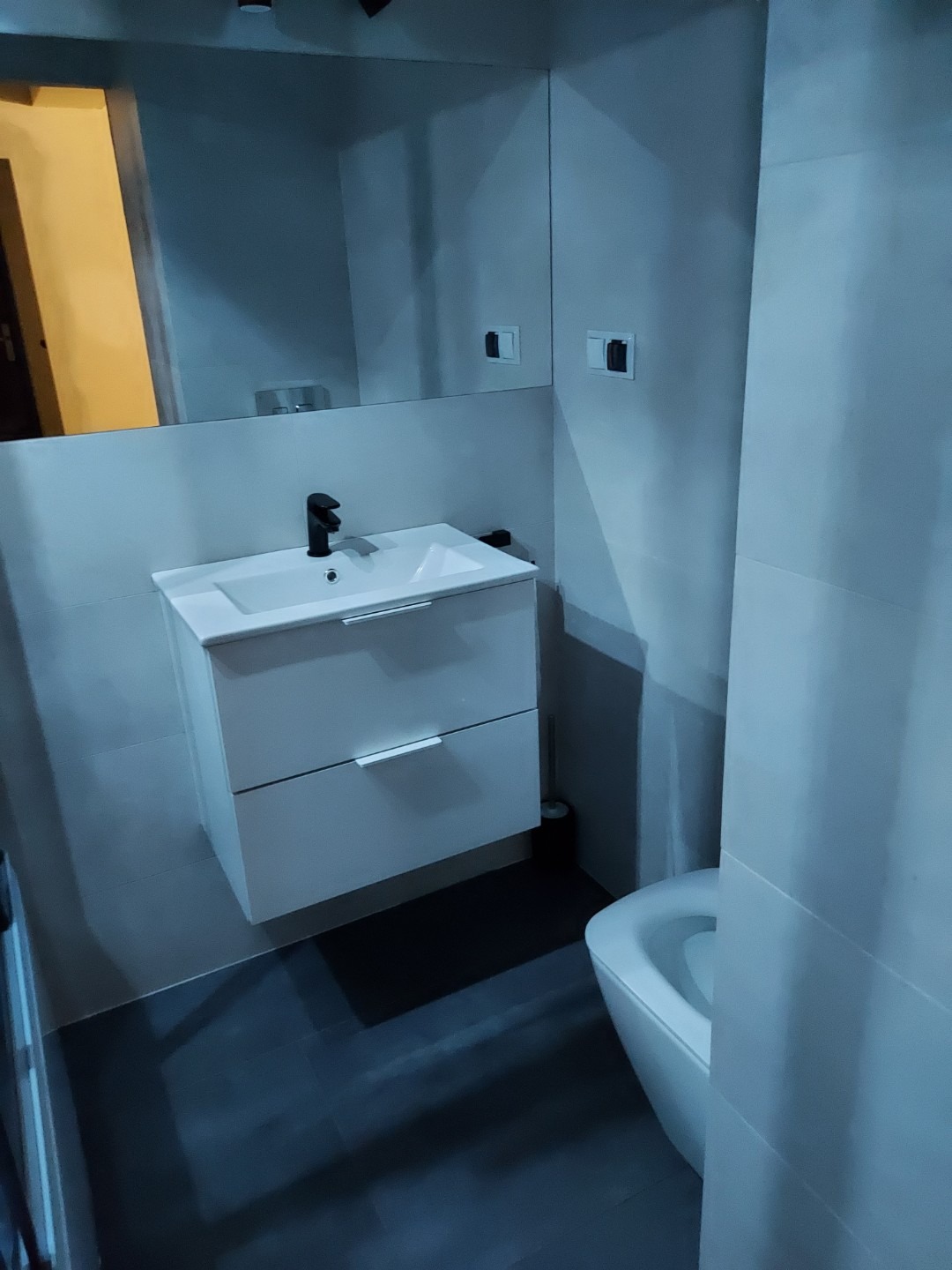 Nowoczesna łazienka z białą szafką pod umywalkę, czarną baterią i dużym lustrem, widok na fragment toalety, szare płytki na ścianach i podłodze.