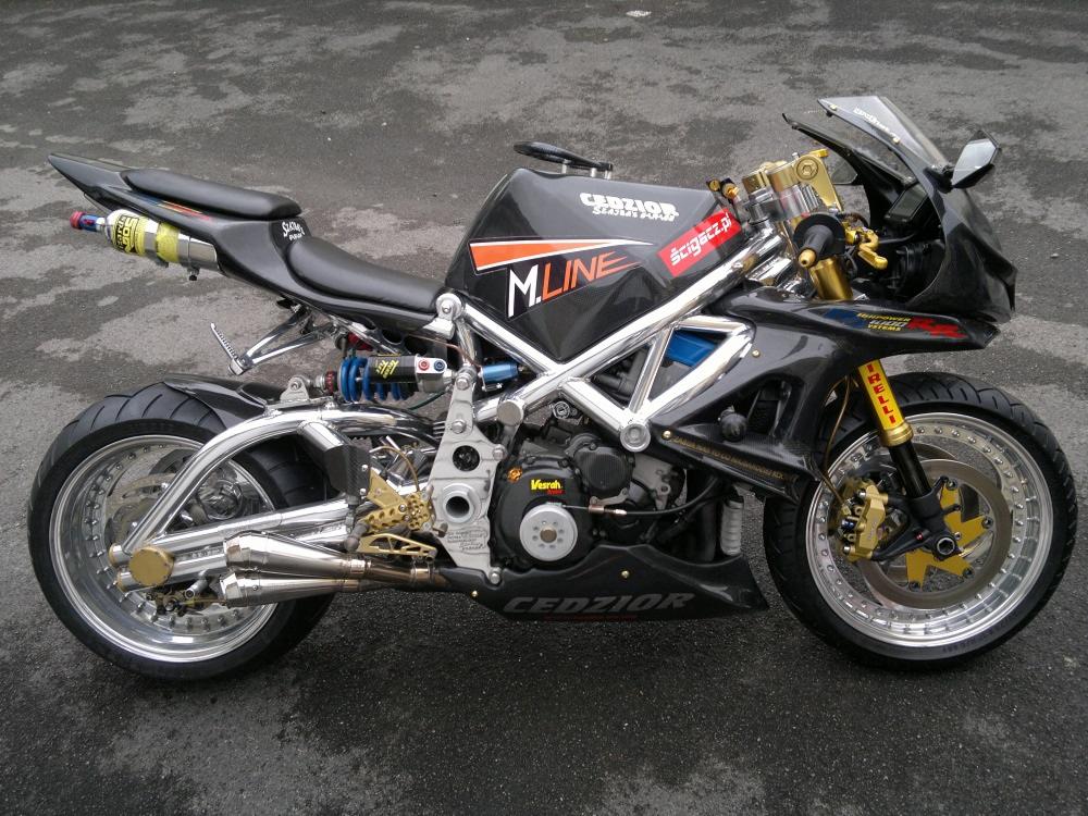 Czarny motocykl typu custom z chromowaną ramą i złotymi akcentami, stojący na szarym asfalcie. Widoczne logo 'CEDZIOR' i naklejki.