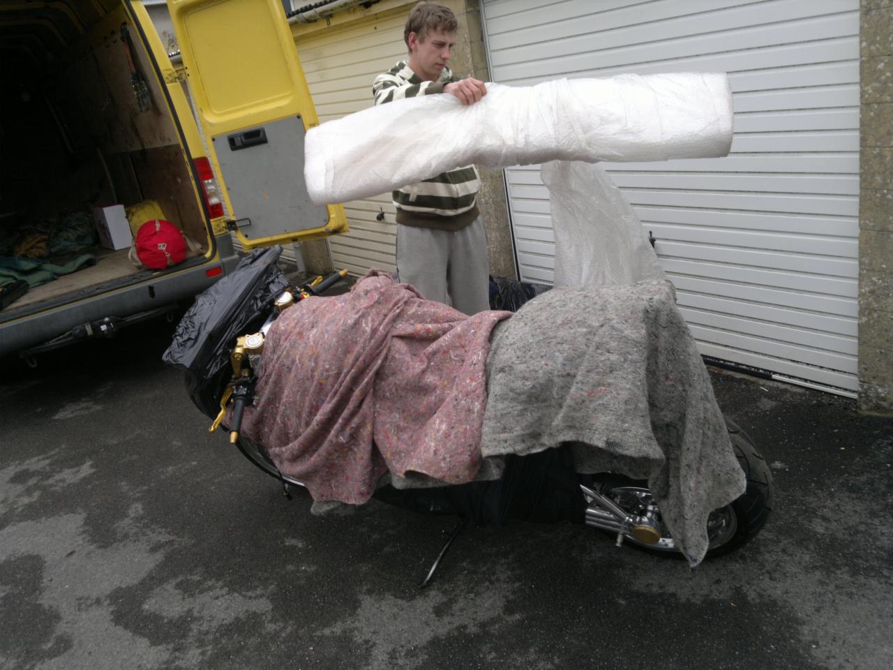 Mężczyzna zabezpiecza motocykl folią i kocami przed transportem w żółtym busie. Widoczne otwarte drzwi busa i brama garażowa w tle.