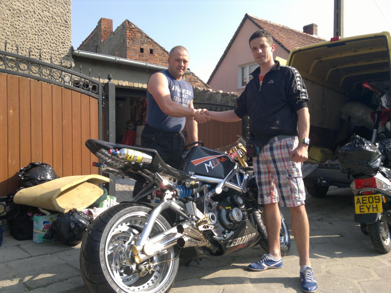 Dwóch mężczyzn w uścisku dłoni obok motocykla i żółtego busa, prawdopodobnie po zakończonym transporcie. Widoczne torby i elementy wyposażenia.