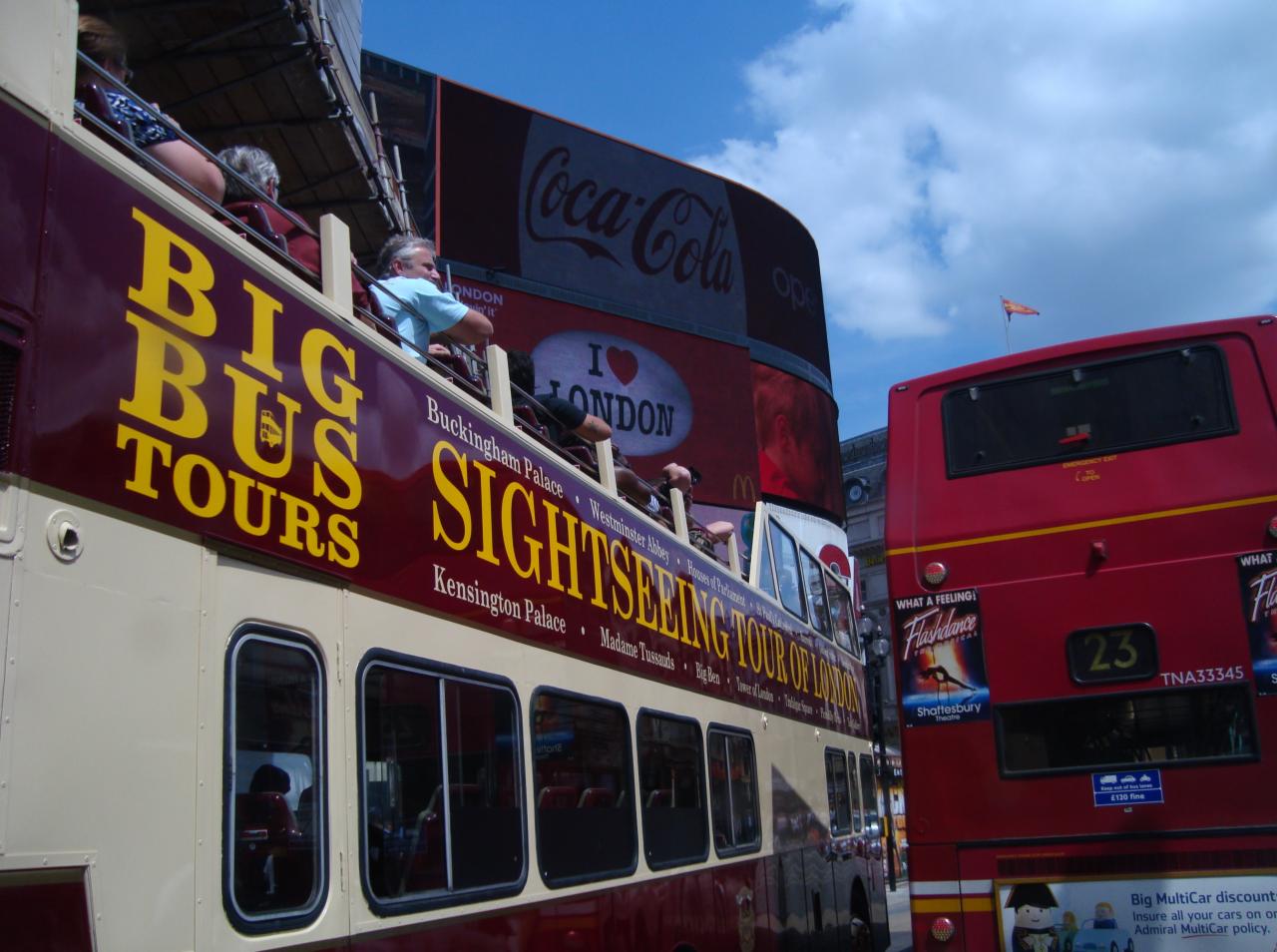 Otwarty autobus turystyczny 'Big Bus Tours' w Londynie, z pasażerami na pokładzie i reklamą Coca-Coli w tle. Widoczny fragment drugiego autobusu.