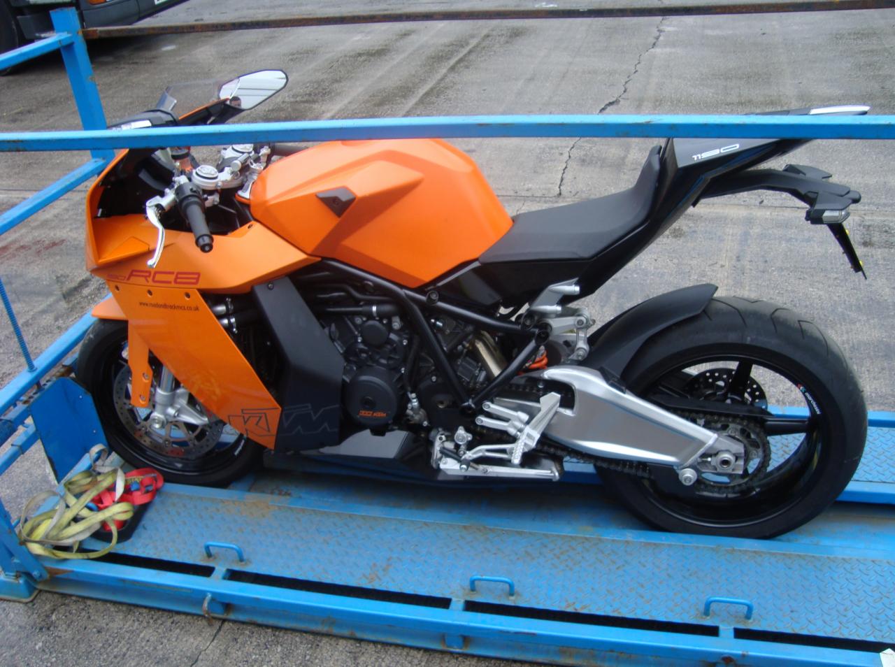 Pomarańczowy motocykl KTM 1190 RC8 na niebieskiej platformie transportowej, widok z boku, lekkie pochylenie w lewo.