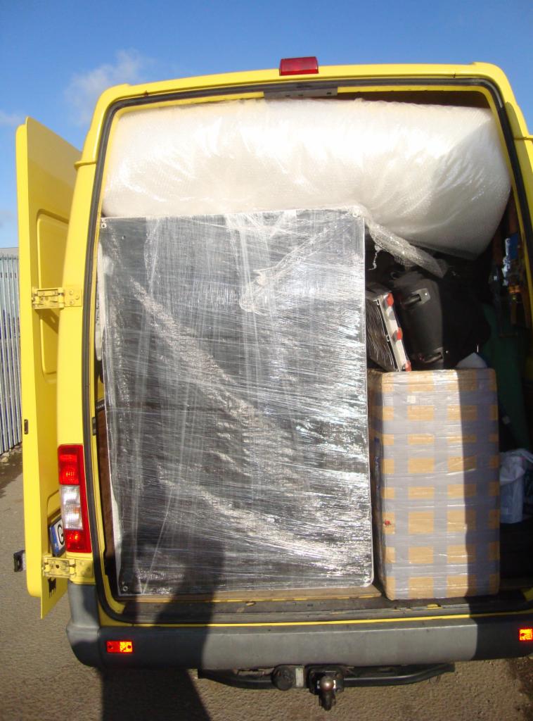 Żółty bus z otwartymi drzwiami, załadowany meblami owiniętymi folią, gotowy do transportu. Widok z tyłu pojazdu.