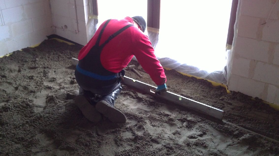 Pracownik w czerwonej bluzie i ogrodniczkach wyrównuje mokrą, szarą wylewkę poziomnicą w pomieszczeniu z oknem, widoczne cegły i żółta folia ochronna.