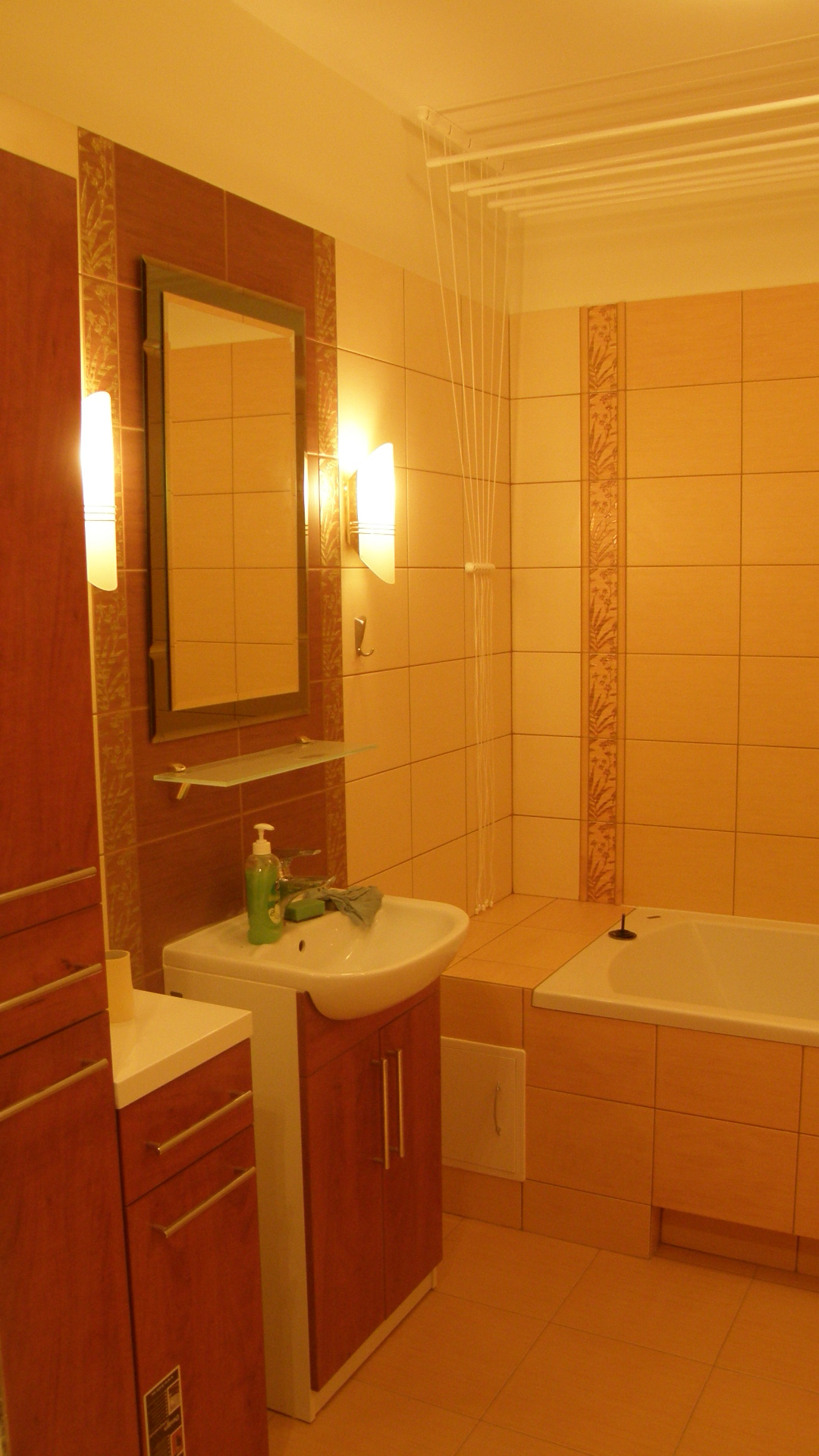 Umeblowana łazienka z brązowymi płytkami, lustrem, umywalką z szafką i suszarką na pranie pod sufitem.