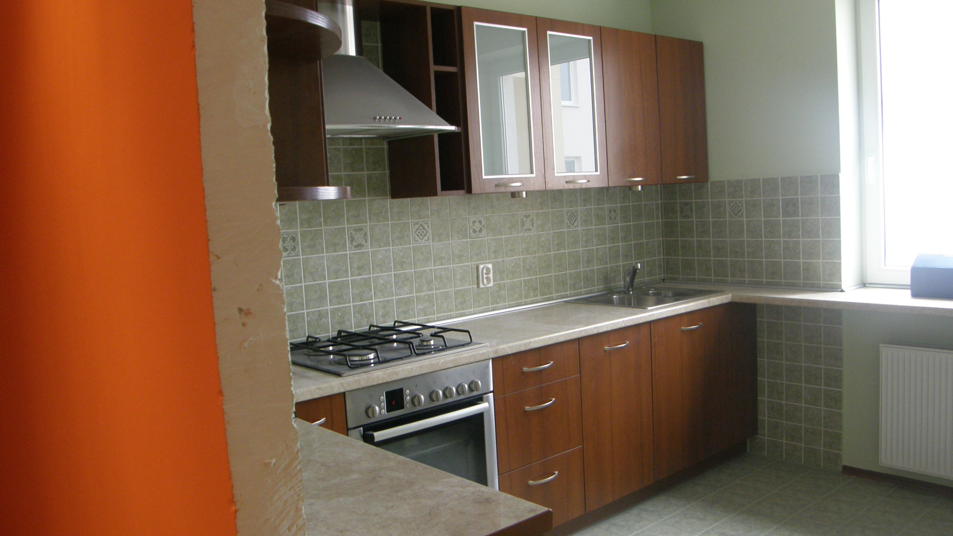 Kuchnia po remoncie: zabudowa z drewnianymi szafkami, płytki ścienne w odcieniach szarości i beżu, widoczny fragment pomarańczowej ściany przed wykończeniem.