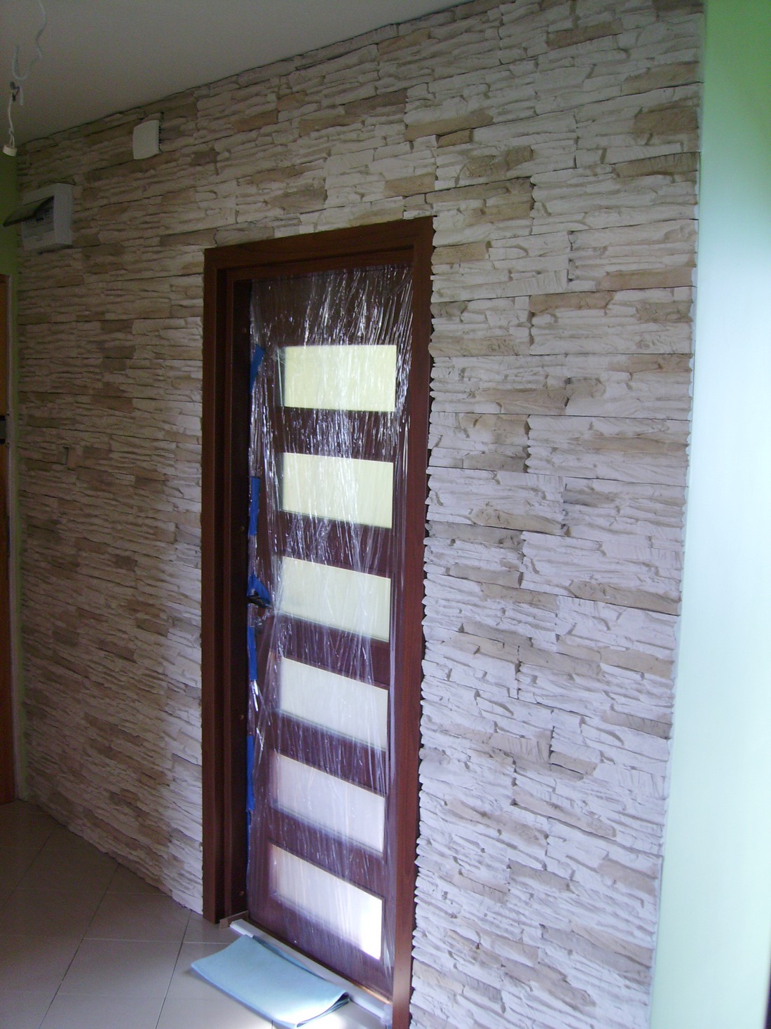 Ściana wykończona kamieniem dekoracyjnym, z zabezpieczonym folią nowym skrzydłem drzwiowym z przeszkleniami, przed dalszymi pracami wykończeniowymi.