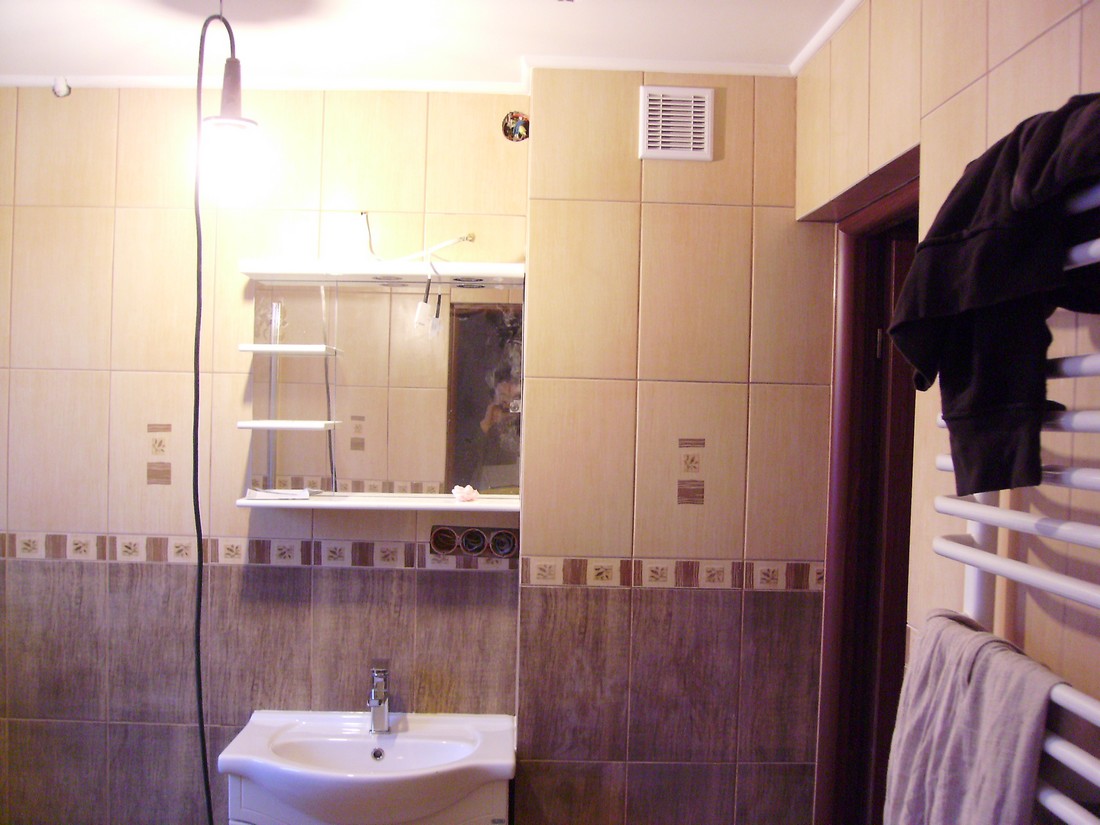 Umywalka z szafką i lustrem w łazience wyłożonej beżowymi i brązowymi płytkami, widoczny grzejnik drabinkowy z powieszonymi ubraniami i prowizoryczne oświetlenie.